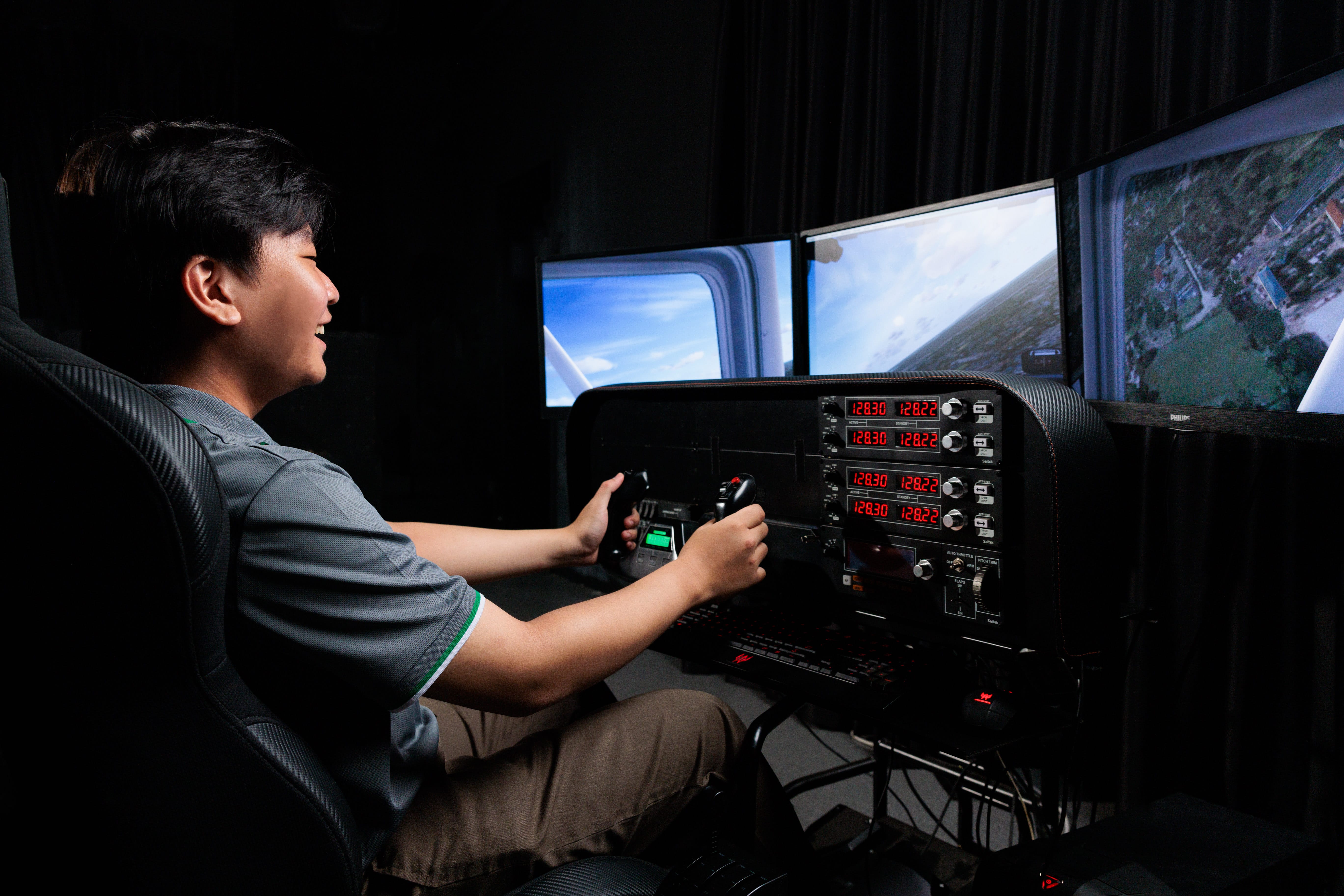 A smiling young man sits at a flight simulator with a joystick, controls, and multiple screens showing sky and land views.